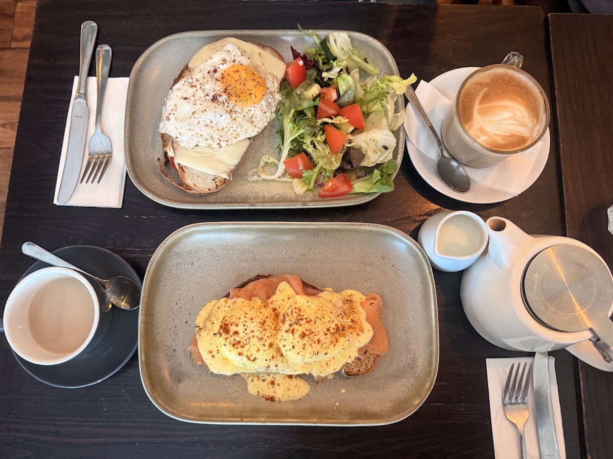 A toasted cheese sandwich topped with a fried egg beside a dressed side salad, with a latte and a teapot.